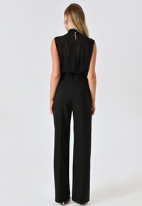 Combinaison noire avec un corsage transparent, un col montant et un pantalon évasé. Présente une fermeture à boutons dans le dos et une texture lisse.