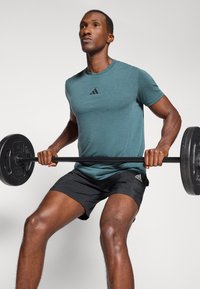 Camiseta deportiva color azul verdoso con un logo negro, combinada con pantalones cortos negros. La persona levanta una barra con pesos circulares. Fondo simple.