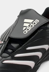 Scarpa sportiva nera con tomaia in pelle sintetica, caratterizzata da accenti bianchi, pannelli laterali testurizzati e un logo in rilievo sulla linguetta.