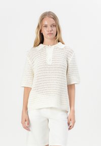 Sweter polo dzianinowy w kolorze kremowym, z krótkimi rękawami, kołnierzykiem oraz luźnym krojem z otwartym splotem i detalami w postaci guzików.