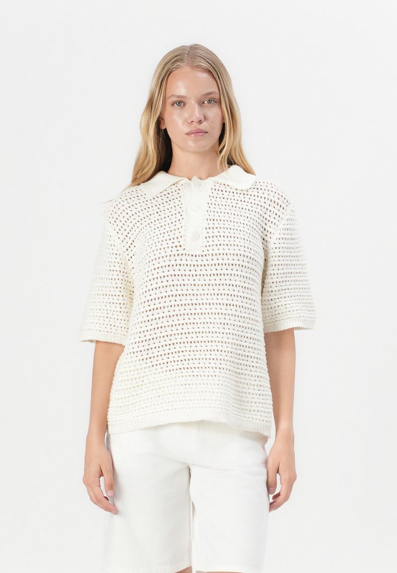 Sweter polo dzianinowy w kolorze kremowym, z krótkimi rękawami, kołnierzykiem oraz luźnym krojem z otwartym splotem i detalami w postaci guzików.