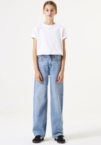 Camiseta blanca de algodón con mangas enrolladas, combinada con unos vaqueros de pierna ancha de color azul claro. Zapatillas negras completan el atuendo.