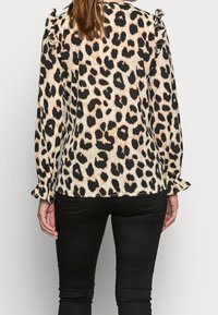 Blouse à imprimé léopard avec des épaules à volants, des manches longues et une coupe ample. Le fond est beige avec des taches noires, associée à un pantalon noir.