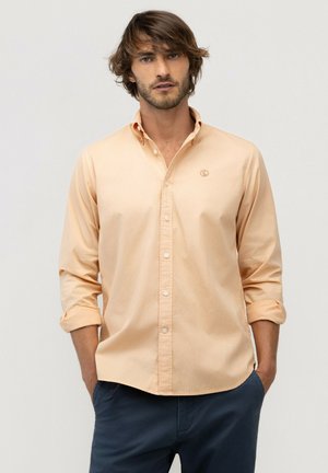 Hombre con cabello castaño de longitud media, vestido con una camisa color melocotón abotonada con las mangas remangadas y pantalones azul oscuro, de pie con las manos en los bolsillos.
