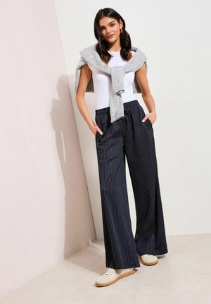 Woman wearing white sleeveless top, gray sweater tied around shoulders, loose black pinstripe pants, and white sneakers, standing against a wall.