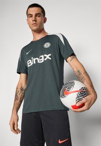 Chelsea-tema atletisk skjorte i mørkegrå med hvide accenter, med et logo og store 'Bingx'-tekster. Modellen holder en mønstret fodbold.