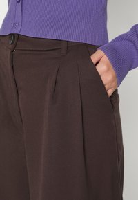 Main dans la poche d'un pantalon plissé marron foncé, portant un pull à manches longues côtelé violet avec des détails de boutons.