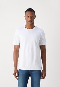 White cotton t-shirt with short sleeves, a round neck, and a small logo detail on the chest, paired with blue jeans. Simple design.