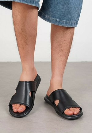Personne portant des sandales noires ouvertes à bout ouvert avec un design ajouré, associées à un short en jean longueur genou sur un sol gris.