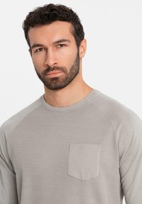 Uomo con capelli corti e ricci di colore scuro e barba, che indossa una camicia a maniche lunghe grigia chiara con taschino, su uno sfondo bianco semplice.