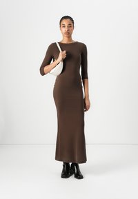 Brown ribbed long-sleeve dress with a fitted silhouette, paired with a white handbag and black ankle boots. Smooth texture and simple design.