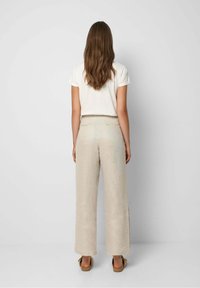 Kvinna med långt brunt hår iförd en vit kortärmad topp, beige vida byxor och bruna sandaler som står framför en enhetlig ljus bakgrund.