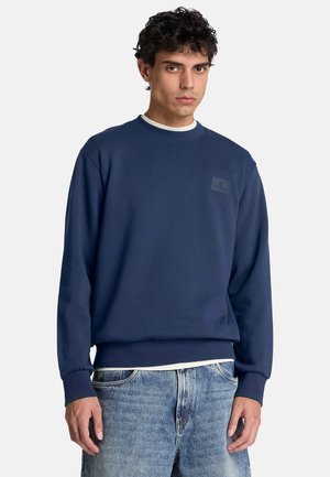 Man draagt een marineblauwe trui met geribbelde manchetten en zoom, blauwe jeans en een wit overhemd zichtbaar bij de kraag en taille.