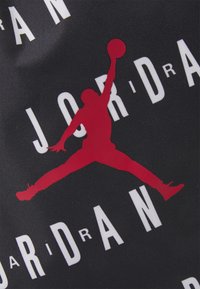 Crni materijal s velikim crvenim siluetom skakača, uz riječ "JORDAN" u podebljanom bijelom slovu.