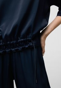 Navyblaues Satin-Oberteil mit geraffter Taille. Verfügt über ein gerüschtes Elastikband und Schnürdetail, mit leichtem, glattem Stoffspiel.