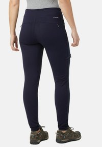 Pantaloni elasticizzati blu navy con vita alta, con tasche laterali con zip e una texture liscia. Abbinati a scarpe da trekking marroni.