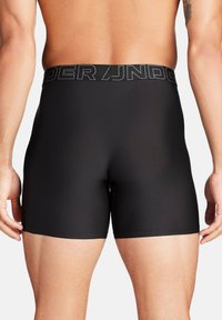 Schwarze Herren Boxershorts aus einem glatten, dehnbaren Material mit einem breiten elastischen Bund, auf dem "UNDER ARMOUR" in grauer Schrift steht.