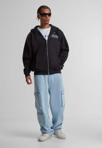 Black zip-up hoodie with large front pockets, paired with light blue cargo pants featuring large side pockets and white sneakers.
