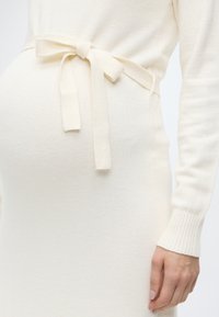 Πλεκτό κρεμ φόρεμα με μακριά μανίκια, με εφαρμοστή μέση και λεπτομέρεια φιόγκου. Απαλή υφή και ριμπ μανσέτες ολοκληρώνουν το σχέδιο.