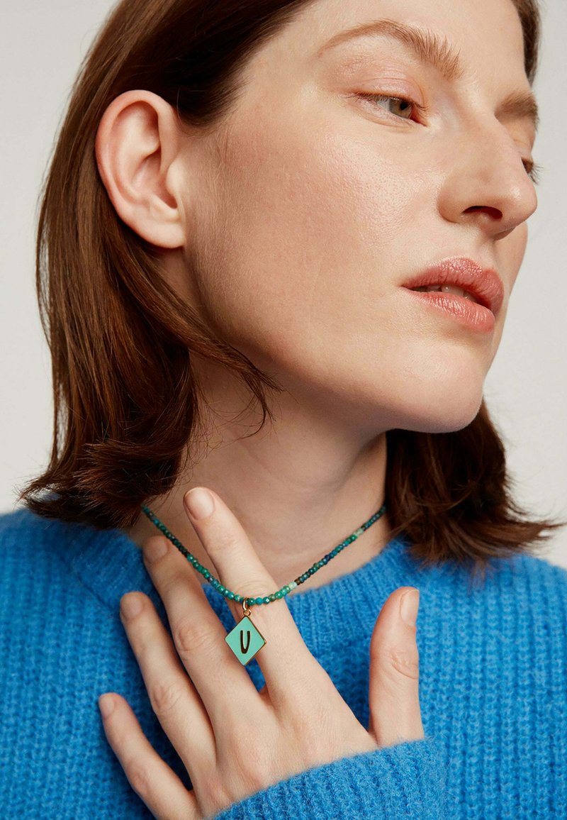 Collar de cuentas en tono verde azulado con un colgante en forma de diamante que presenta una 'U' negra. El modelo lleva un suéter de punto azul, mostrando el collar de manera prominente.