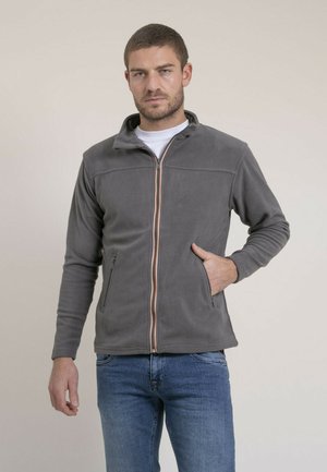 Homme portant une veste polaire grise zippée, une main dans la poche, et un jean bleu sur un fond uni.