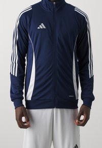 Marineblå träningsjacka med vita ränder, hög krage, hel dragkedja och "AEROREADY"-märke. Tillverkad av glatt, lätt tyg.