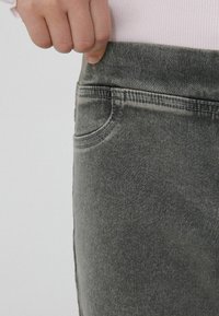 Szare dżinsy z efektem sprania, z boczną kieszenią i gładkim pasem. Zbliżenie na obszar pasa i szczegóły szwów.