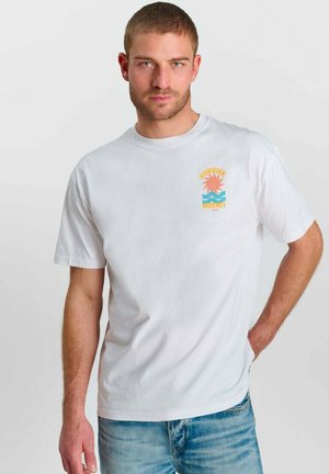 Man draagt een wit T-shirt met korte mouwen met een oranje zon en blauwe golven afbeelding, gecombineerd met lichtblauwe spijkerbroek, staand tegen een effen achtergrond.