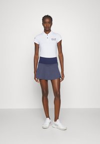 Polo bianco a maniche corte, cintura blu navy, gonna a fantasia e scarpe da ginnastica bianche. Logo EA7 sulla maglietta, texture liscia.