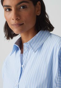 Camicia a righe azzurro chiaro con colletto classico, caratterizzata da bottoni bianchi e una texture di tessuto morbida. Primo piano del colletto e dello scollo.