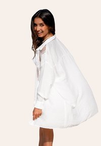 Biała, oversize sukienka koszulowa z zapięciem na guziki z przodu, długimi rękawami i siateczkowymi detalami na ramionach. Teksturowany materiał. Modelka uśmiechająca się.