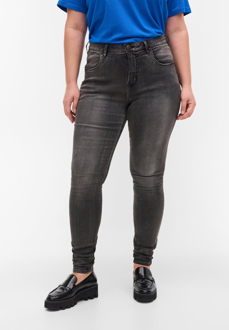 Jeans skinny en denim gris foncé, avec une taille haute, une texture délavée, des poches avant et une fermeture à bouton. Portés avec des mocassins noirs.