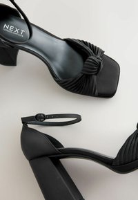 Sorte højhælede sandaler med firkantet tå, knude-detalje på overdelen og en kraftig hæl; justerbar ankelrem. Glat tekstureret finish.