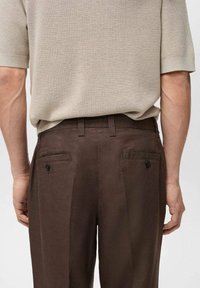 Pantalon marron avec deux poches arrière, doté de boutons noirs. Tissu texturé, coupe ajustée et silhouette à jambes droites.
