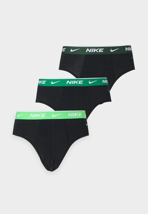 Uomo in piedi con pelle scura che indossa slip Nike neri con fascia verde su sfondo grigio uniforme.