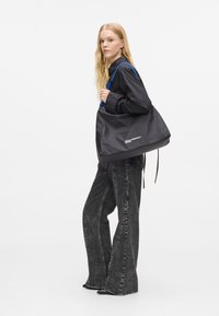 Γυναίκα με μακριά ξανθά μαλλιά φοράει μαύρο πουκάμισο και φαρδιά μαύρα τζιν, κρατώντας μεγάλη μαύρη τσάντα ώμου με μπλε λουριά.