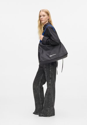 Γυναίκα με μακριά ξανθά μαλλιά φοράει μαύρο πουκάμισο και φαρδιά μαύρα τζιν, κρατώντας μεγάλη μαύρη τσάντα ώμου με μπλε λουριά.