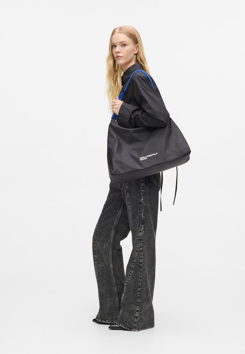 Γυναίκα με μακριά ξανθά μαλλιά φοράει μαύρο πουκάμισο και φαρδιά μαύρα τζιν, κρατώντας μεγάλη μαύρη τσάντα ώμου με μπλε λουριά.