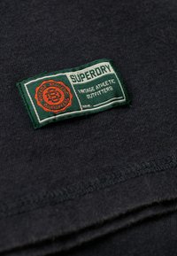 Sort bomulds T-shirt med et grønt og hvidt mærke, der har "Superdry" og "Vintage Athletic Outfitters" i orange og hvid tekst.
