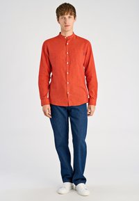 Camicia rossa a maniche lunghe con collo abbottonato e taschino; abbinata a jeans blu scuro e sneakers bianche. Tessuto liscio, vestibilità casual.