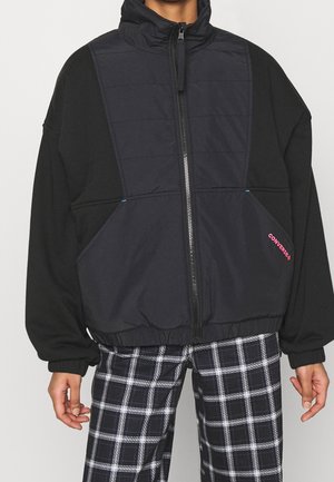 Persoon draagt zwarte gewatteerde bodywarmer met rits en zijzakken, over zwarte mouwen en zwart-witte geruite broek.