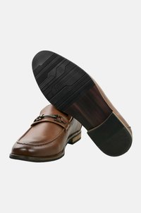 Mocasines de cuero marrón con punta redondeada, suela de goma negra, acento metálico decorativo y detalles de costura a lo largo del borde superior.