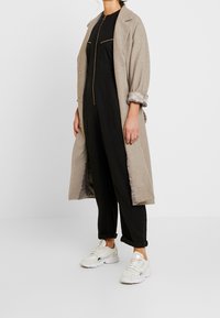 Beige lange jas over een zwarte jumpsuit, met ritszakken en een relaxte pasvorm, gecombineerd met witte sneakers, waarmee textuurcontrasten worden benadrukt.