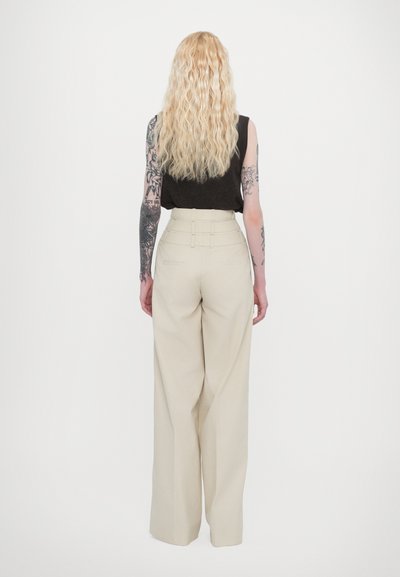 Beige wide-leg trousers with high-waisted design, side pockets, and belt loops. Model wears a black sleeveless top and has wavy hair.