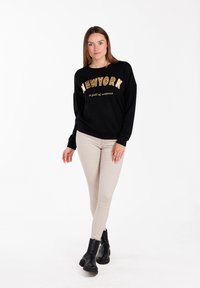 Sudadera negra con la frase "NUEVA YORK" en una tipografía de leopardo dorada, combinada con pantalones skinny beige y botas de tobillo negras. Diseño simple y colores sólidos.