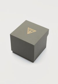 Boîte carrée grise à surface texturée avec le logo Guess doré sur le couvercle, posée sur un fond blanc.