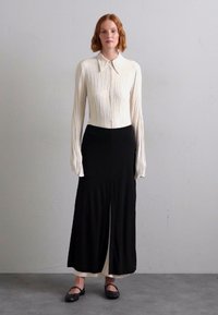 White, pleated long-sleeve blouse paired with high-waisted black wide-leg trousers and black flat shoes, set against a neutral background.