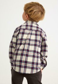 Mały chłopiec z krótkimi blond włosami, ubrany w koszulę w kolorze kremowym, czerwonym i niebieskim w kratę oraz ciemne spodnie, stojący tyłem.