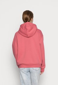 Růžová mikina s kapucí a volným střihem, vyrobená z měkké látky; má šňůrku na kapuci a žebrované manžety, doplněná světle modrými džínami.