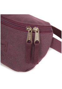 Burgundin värinen vyölaukku, joka on valmistettu teksturoidusta materiaalista. Varustettu vetoketjusulkimella, jossa on kultasävytteinen metalliosat ja kaksi kangasvedintä.